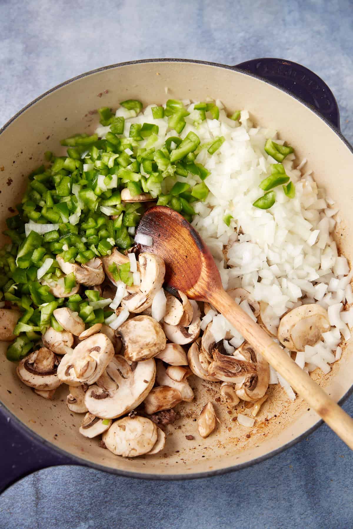 A large pot contains chopped green bell peppers, diced onions, and sliced mushrooms being sautéed, the essential vegetables for a delicious cheesesteak casserole. A wooden spoon rests inside the pot, showing the cooking process in its early stages. The background is a light-colored countertop.