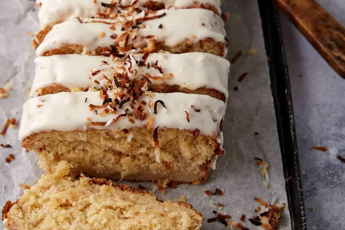 A close-up of sliced coconut loaf cake, a delightful addition to any quick breads collection, topped with white icing and toasted shredded coconut, displayed on parchment paper. One slice is slightly pulled away from the loaf.