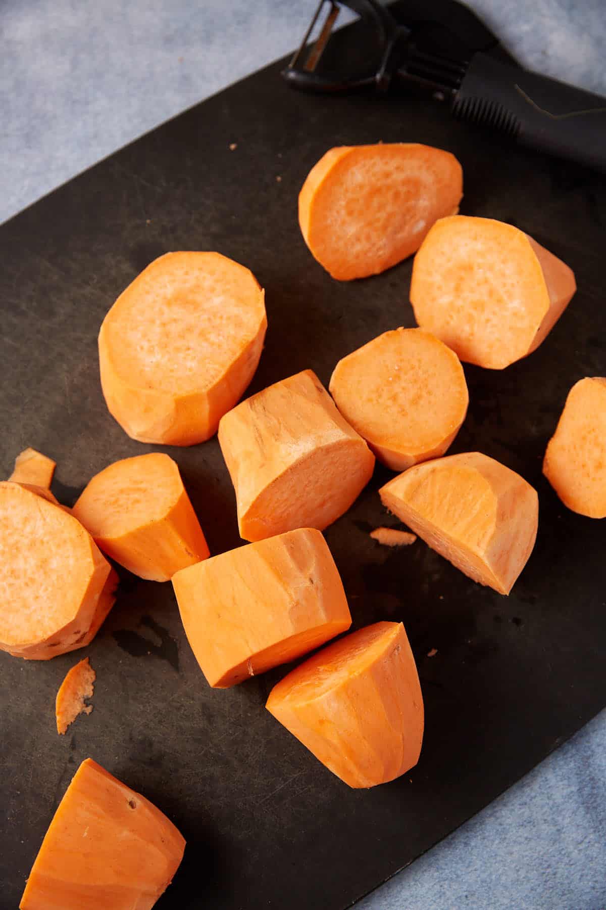 Several pieces of chopped sweet potato are scattered on a black cutting board, ready for the skillet. A peeler rests nearby on the board. The background is light-colored.