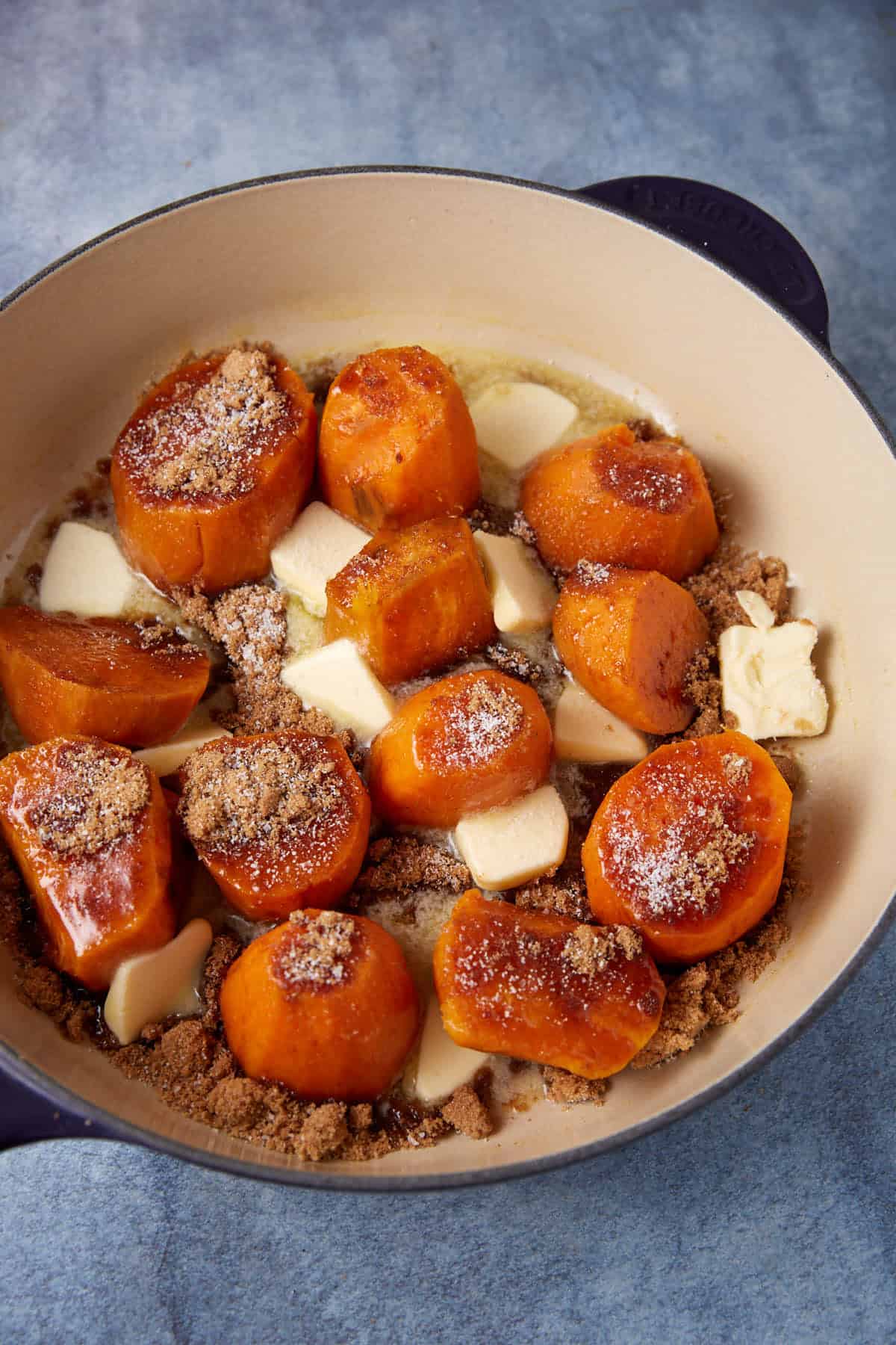 A cooking dish filled with round chunks of sweet potato covered in brown sugar and small pats of butter. The dish is placed against a blueish background, and the sweet potatoes are lightly caramelized.