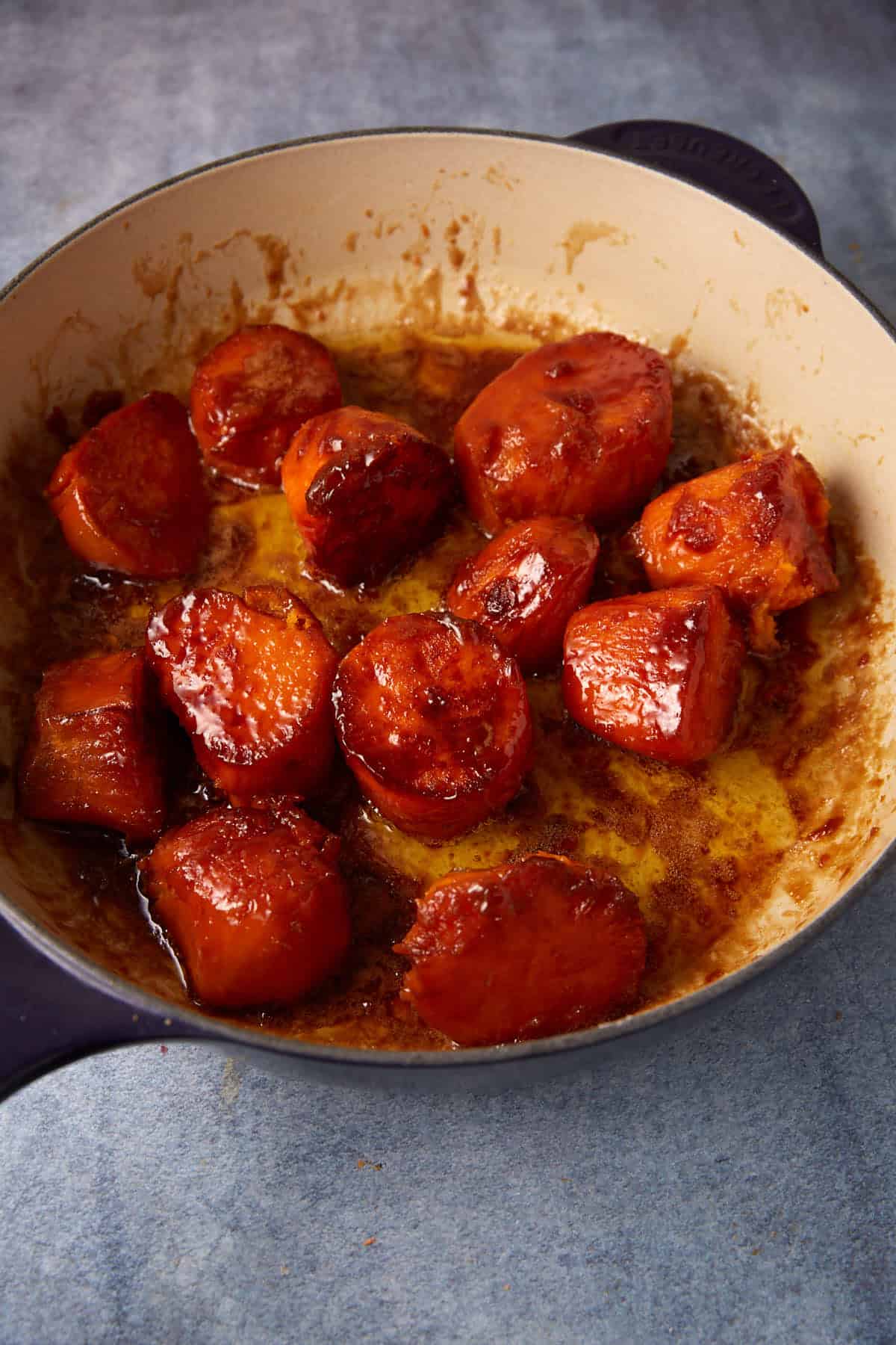 A round baking dish filled with roasted sweet potatoes that are caramelized and glazed. The brightly colored sweet potatoes are cut into thick chunks and appear glossy from the sauce, set on a textured, light-colored surface.