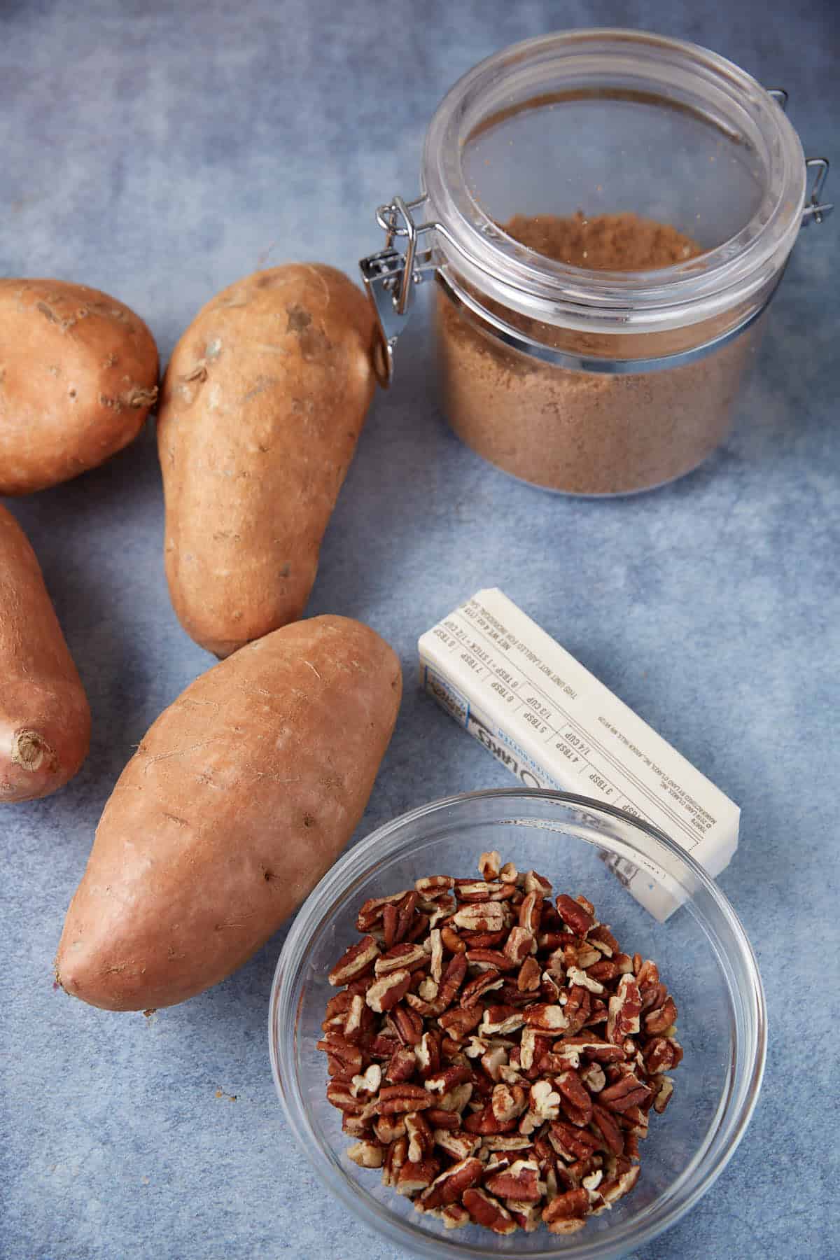 Three sweet potatoes, a stick of butter, a glass jar of brown sugar, and a bowl of chopped pecans are arranged on a blue surface.