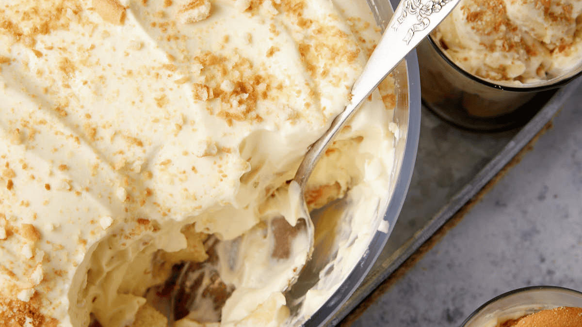 A bowl of creamy, layered banana pudding topped with crushed vanilla wafers, served with a silver spoon. A small glass container of the same dessert is next to it on a gray surface, offering a delightful contrast to your favorite lemon desserts.