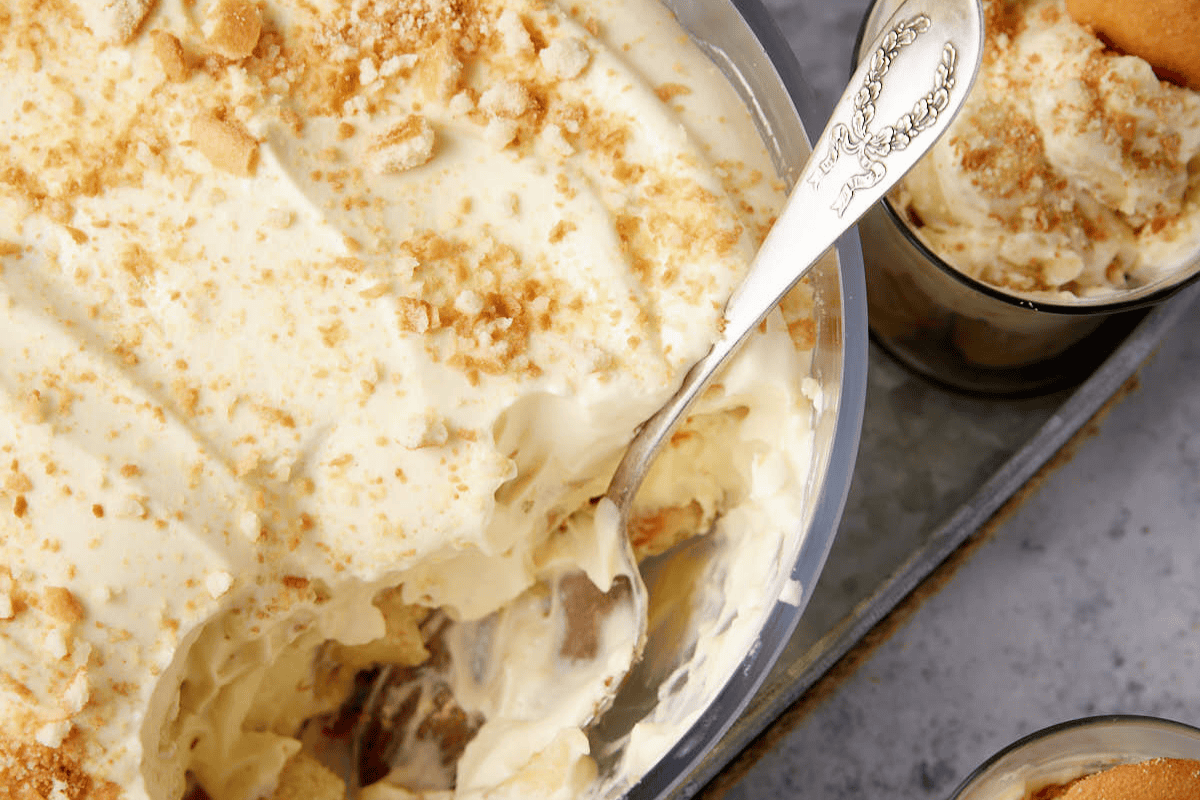 A close-up of a dish filled with creamy banana pudding topped with crushed vanilla wafers. A spoon is partially submerged in the pudding, scooping some into a nearby small bowl, also containing the dessert. The tray and marble background are partially visible.