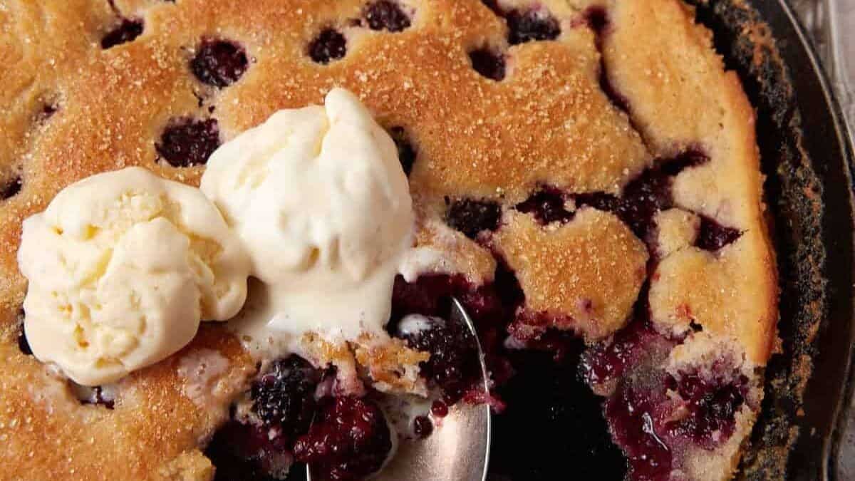 A close-up of a berry cobbler with a golden-brown crust, topped with melting vanilla ice cream scoops. A spoon has scooped out a portion, revealing the juicy berry filling inside.