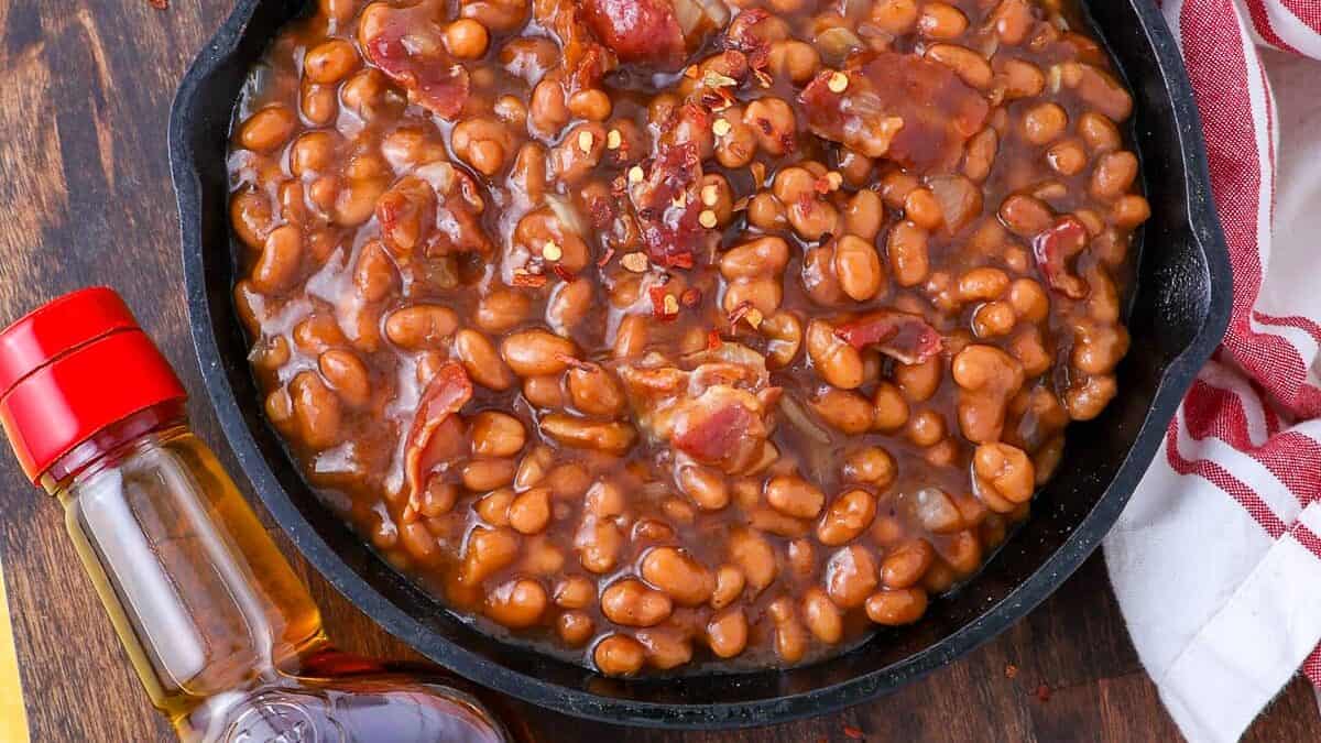 A cast iron skillet filled with baked beans and bacon is placed on a table with a red and white striped cloth. A bottle of Maker's Mark Kentucky Bourbon is positioned nearby.