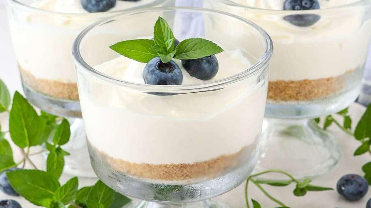 Three glass dessert cups filled with cheesecake, garnished with fresh blueberries and mint leaves. A layer of graham cracker crust is visible at the bottom. The glasses are placed on a white plate decorated with more blueberries and mint sprigs.