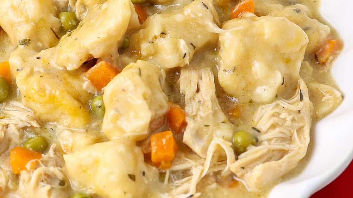 A close-up of a plate filled with a creamy chicken and dumplings dish. Visible ingredients include tender pieces of shredded chicken, fluffy dumplings, carrots, peas, and a thick, seasoned gravy. The meal appears warm and hearty, served in a white bowl.