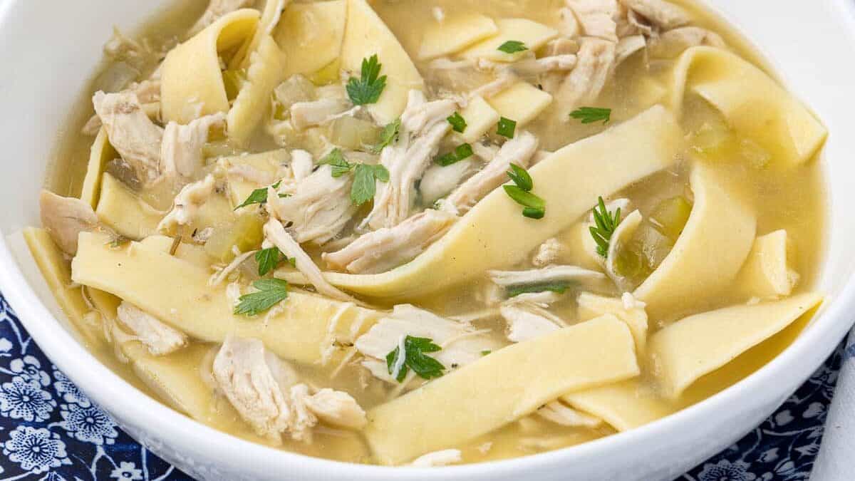 A white bowl filled with chicken noodle soup, featuring wide egg noodles, shredded chicken, and garnished with fresh herbs. The bowl is placed on a blue floral-patterned cloth, with several crackers visible in the background.