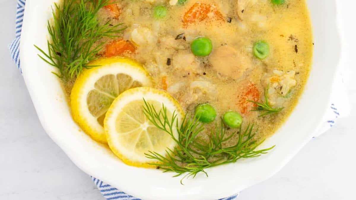 A bowl of creamy chicken soup garnished with peas, carrots, lemon slices, and fresh dill sits on a blue and white patterned cloth. The soup is in a white bowl, and the background is a light marble surface.