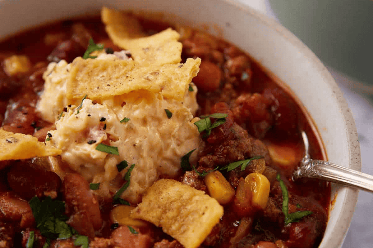 A close-up of a hearty chili dish in a bowl, topped with shredded cheese, corn chips, and finely chopped green herbs. The chili contains kidney beans, corn, and ground meat, creating a rich and colorful appearance. A spoon rests on the edge of the bowl.
