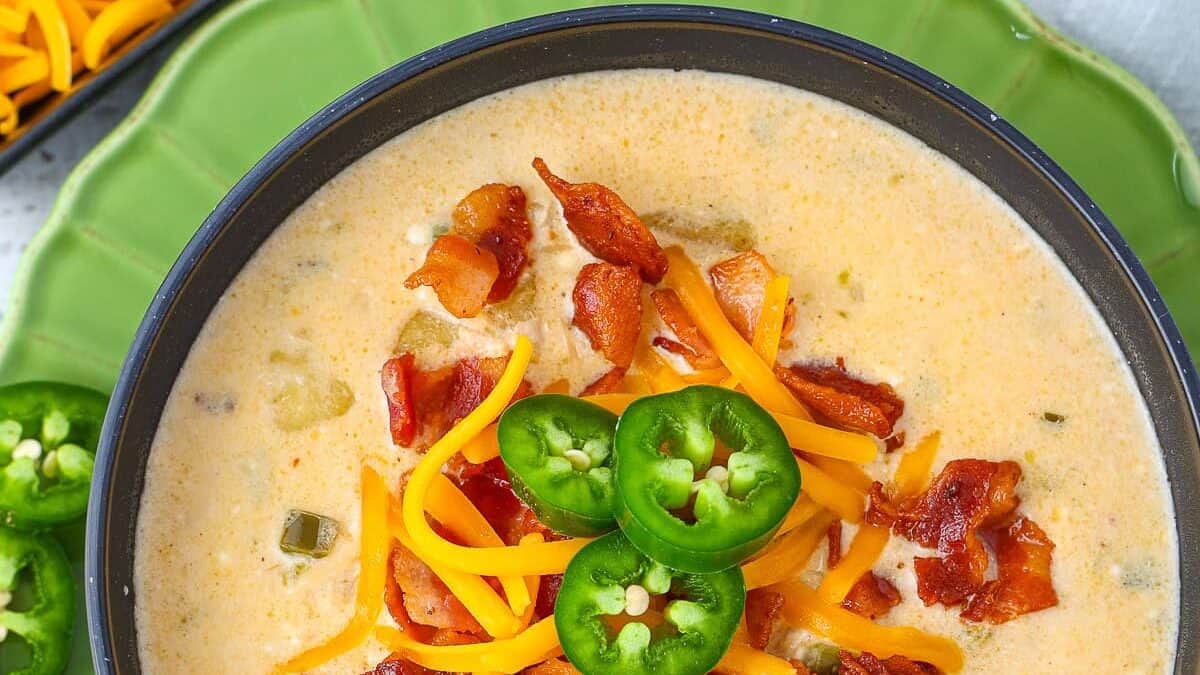 A bowl of creamy white soup garnished with crispy bacon, shredded cheddar cheese, and sliced jalapeños. The bowl is placed on a green plate, with additional sliced jalapeños visible in the background.