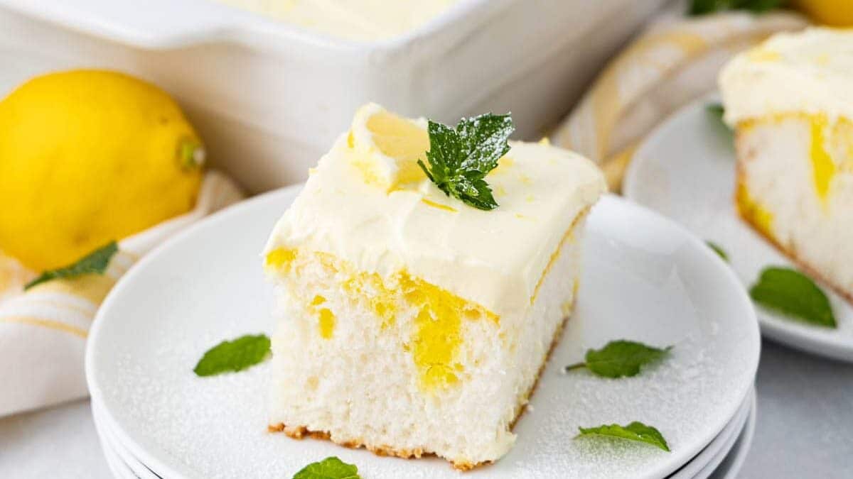 A slice of lemon poke cake with light frosting and a mint garnish sits on a stack of white plates. In the background, a rectangular dish holds the remaining cake, accompanied by a whole lemon and additional mint leaves.