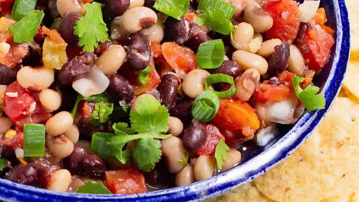 A close-up view of a bowl filled with a colorful bean salad, featuring black beans, black-eyed peas, diced tomatoes, chopped green onions, and fresh cilantro. The bowl is situated next to a pile of tortilla chips on a white surface.