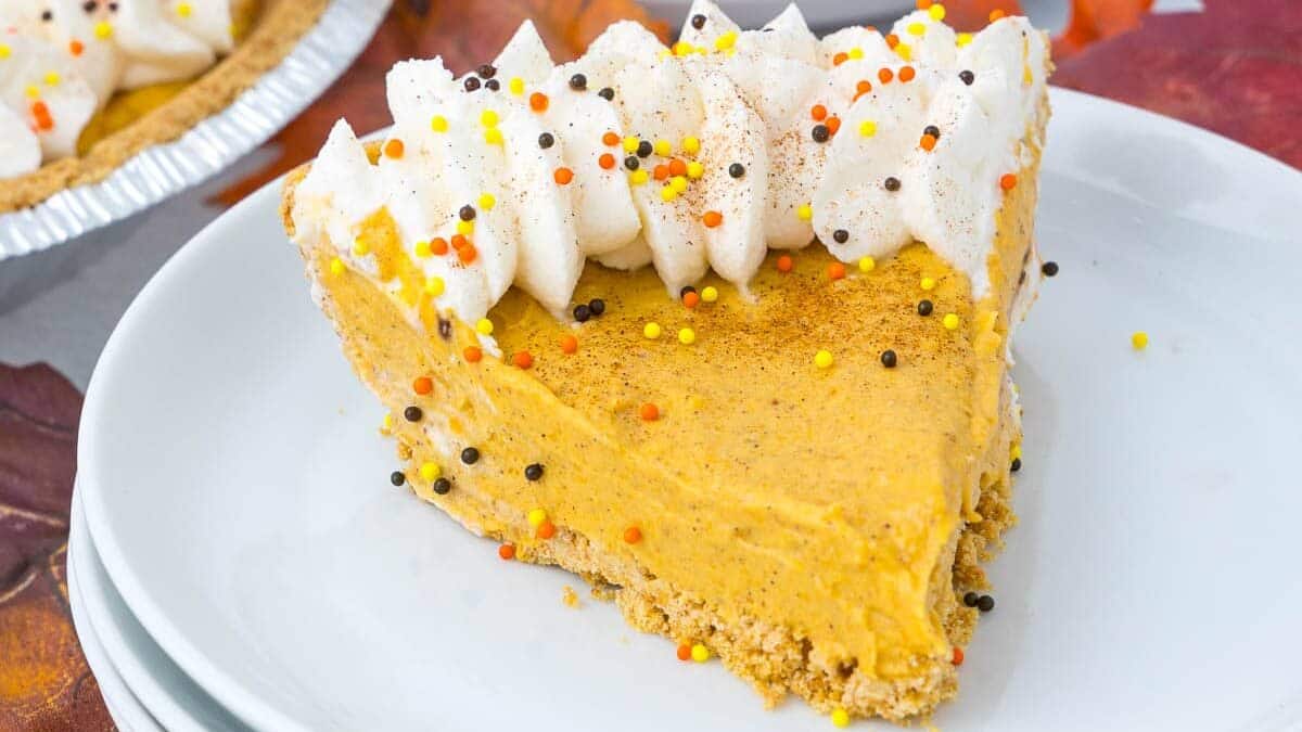 A slice of pumpkin pie topped with whipped cream and colorful sprinkles sits on a stack of white plates. Another partly visible pie and autumn leaves in the background enhance the fall theme.