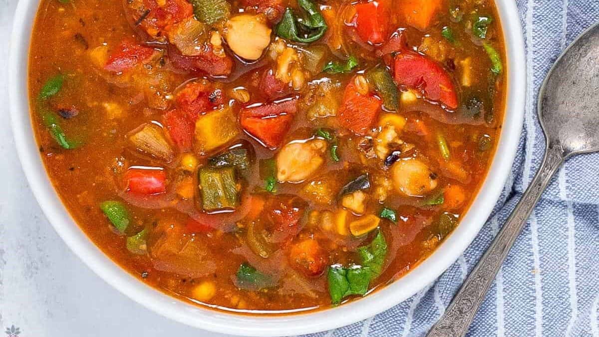 A bowl of vibrant vegetable chili filled with a colorful mix of tomatoes, peppers, zucchini, corn, chickpeas, and spinach, accompanied by a lime wedge and a small dish of hot sauce. A spoon and a striped cloth napkin are placed beside the bowl.