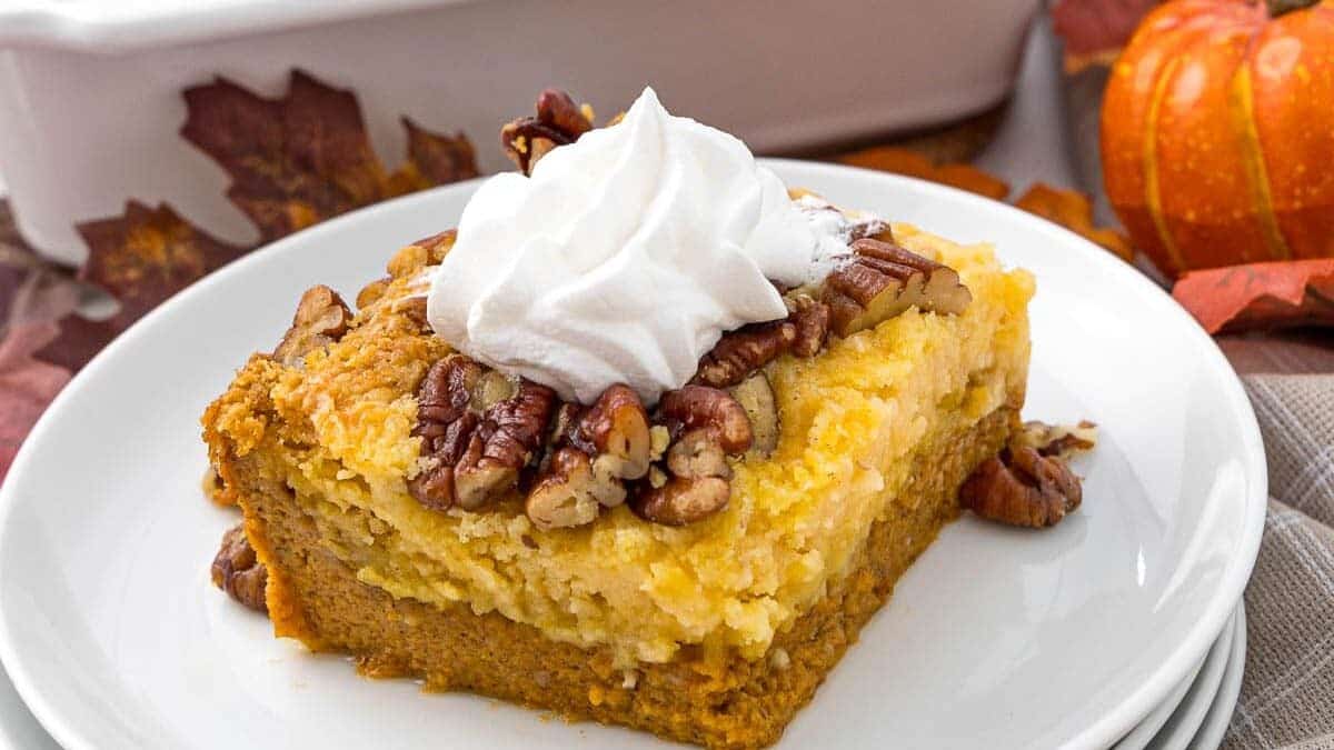 A slice of pumpkin pecan dump cake topped with whipped cream on a stack of white plates. The cake slice has layers of pumpkin, pecans, and yellow cake mix. In the background, a baking dish with more cake and autumn-themed decorations like leaves and a small pumpkin can be seen.