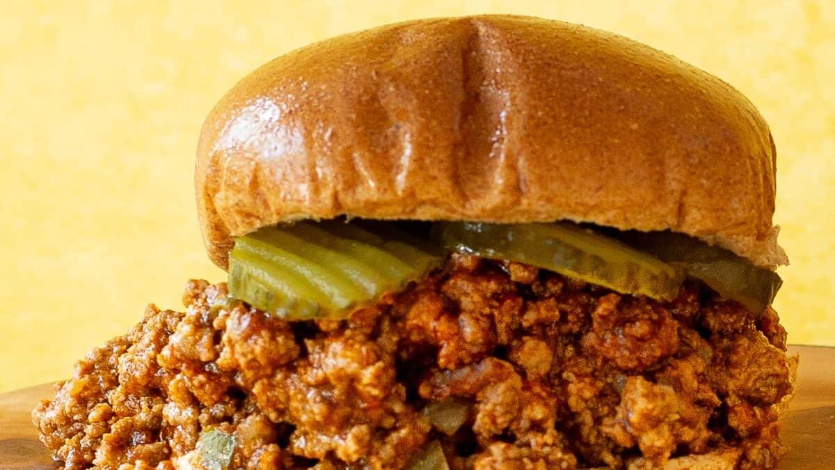 A close-up of a sloppy joe sandwich featuring a generous serving of seasoned ground meat spilling out from a toasted bun, topped with sliced pickles, displayed on a wooden cutting board with a plain yellow background.