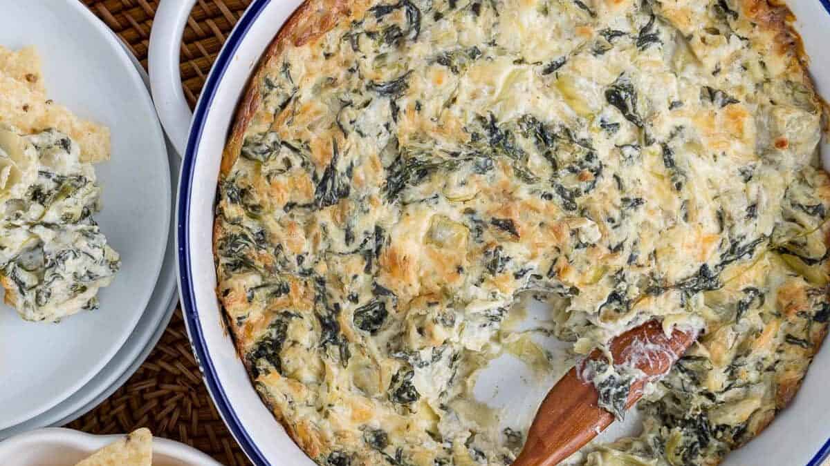 A round white dish filled with creamy spinach and artichoke dip, partially scooped with a wooden spoon. The dish is placed on a wicker mat and accompanied by a plate of tortilla chips, some of which have dip on them. A beige cloth is partially visible underneath.