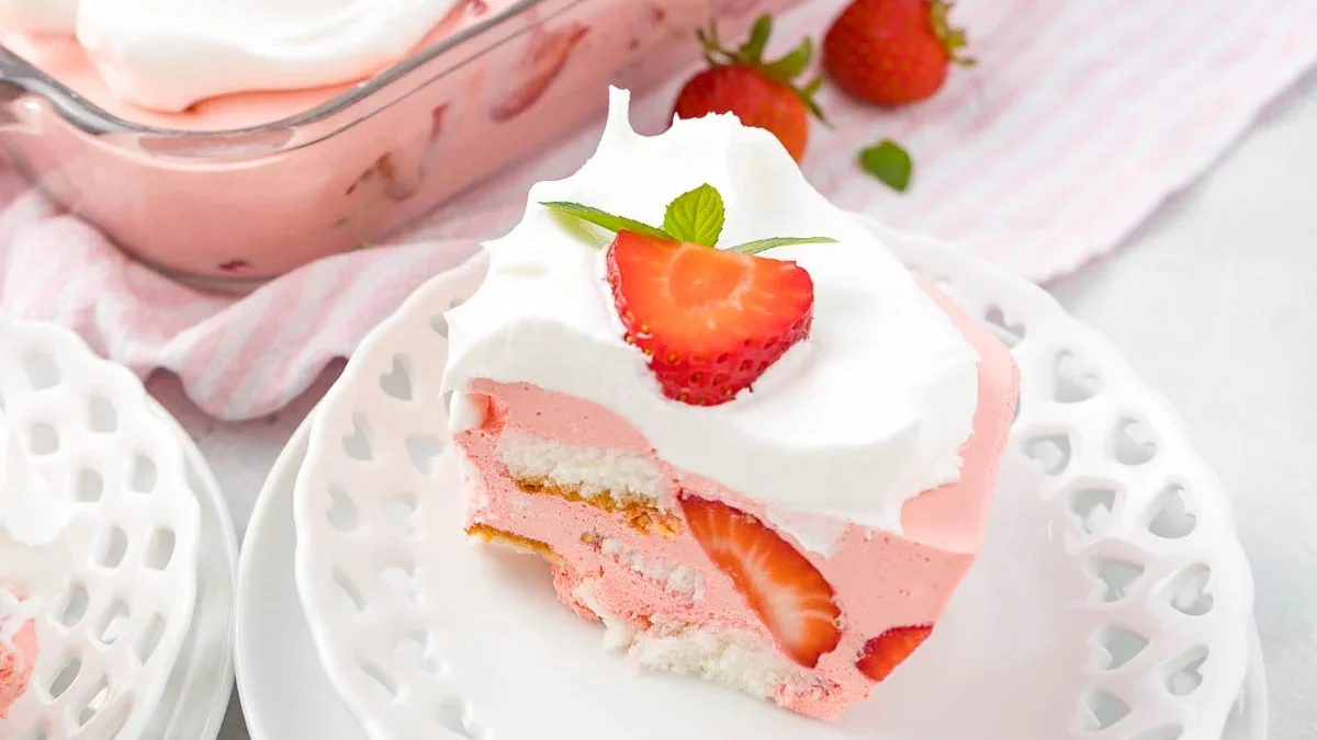 A slice of strawberry icebox cake sits on a decorative white plate. The cake features layers of whipped cream, pink strawberry filling, fresh strawberries, and a biscuit crust. In the background, the rest of the cake is in a glass dish with more strawberries scattered around.
