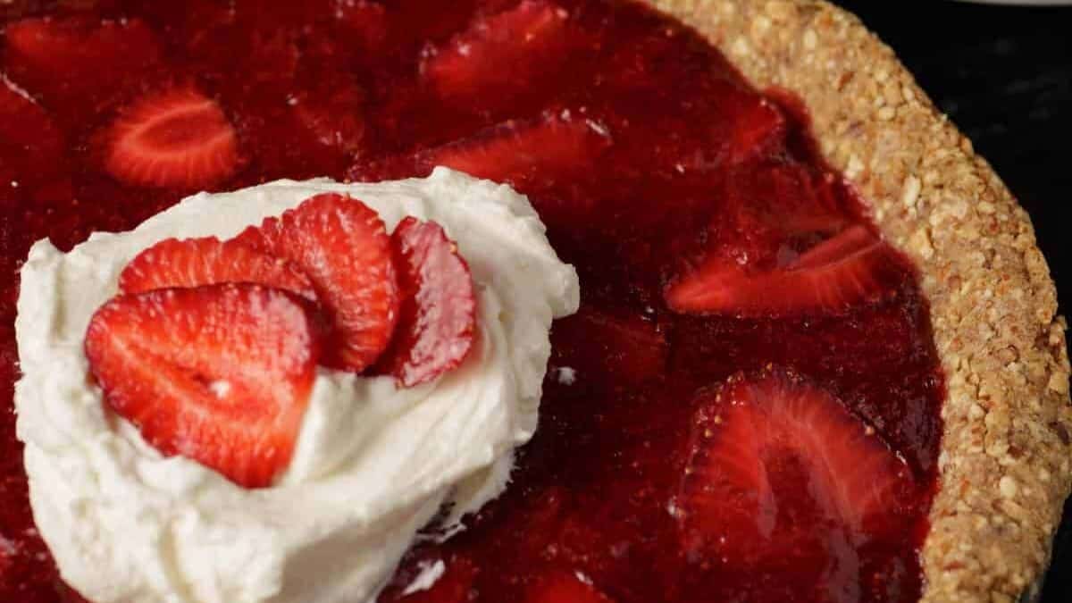 A close-up of a strawberry pie with a golden-brown crust. The filling is made of fresh, bright red strawberry slices encased in a glossy red glaze. The pie is topped with a dollop of whipped cream and garnished with several fresh strawberry slices.