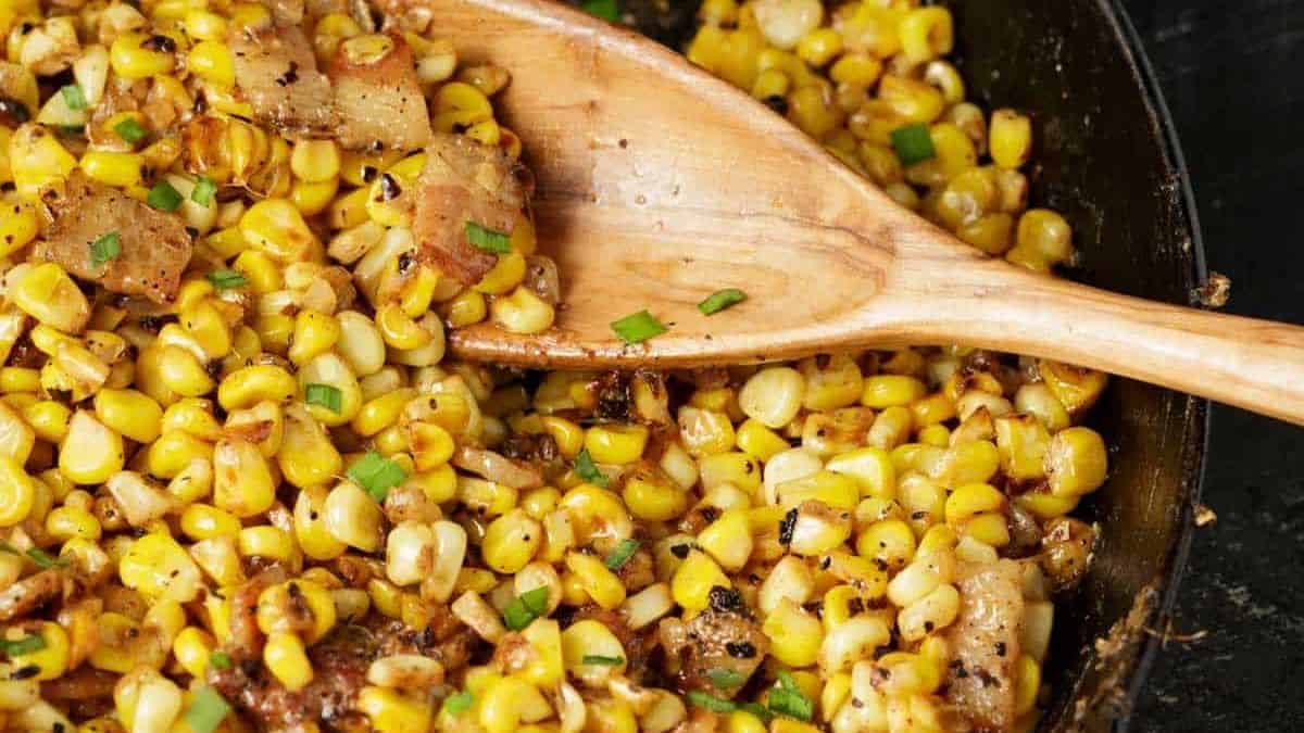 A close-up of a skillet filled with freshly cooked corn sautéed with bits of onions, green herbs, and seasonings. A wooden spoon is stirring the mixture, which appears golden and slightly charred in some spots, highlighting a delicious and inviting texture.
