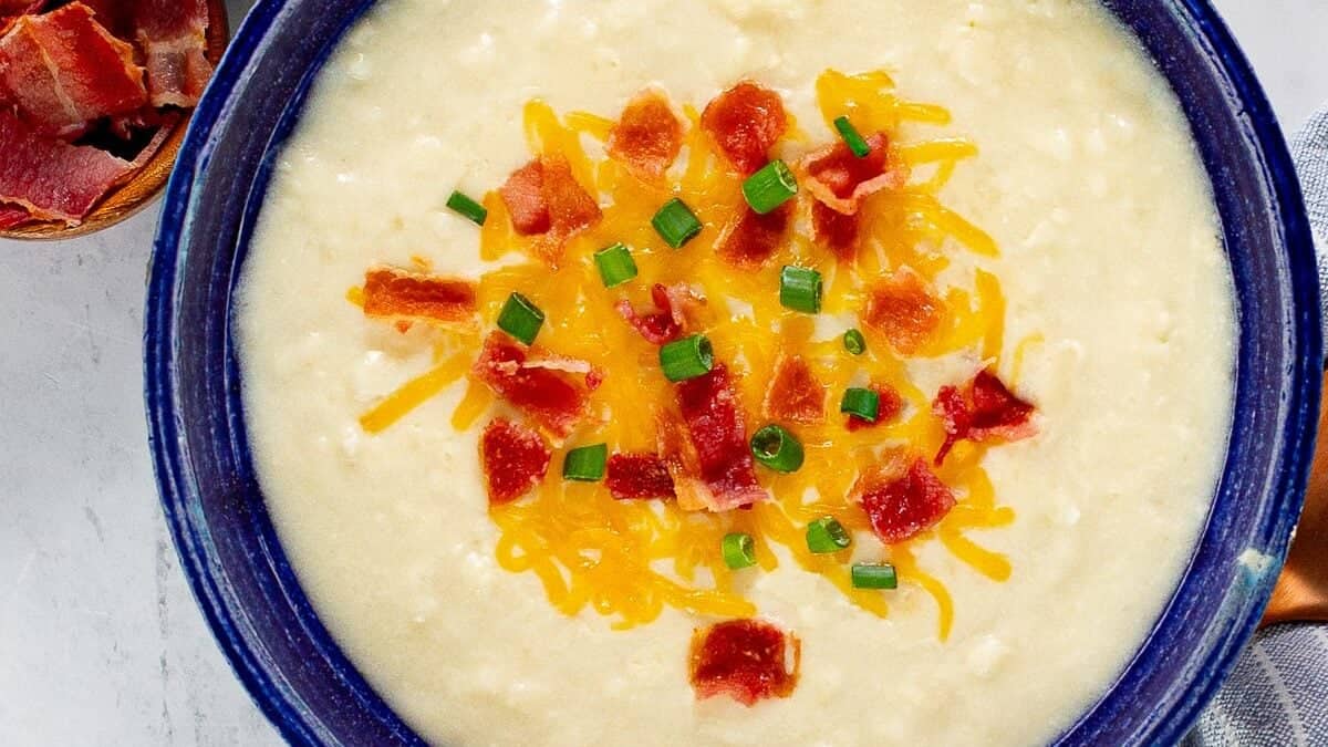 A bowl of creamy potato soup topped with shredded cheese, chopped bacon, and green onions. Beside the bowl are small dishes containing extra shredded cheese and bacon pieces. A blue-striped cloth napkin and a wooden spoon are next to the bowl—a perfect example of comforting potato recipes.