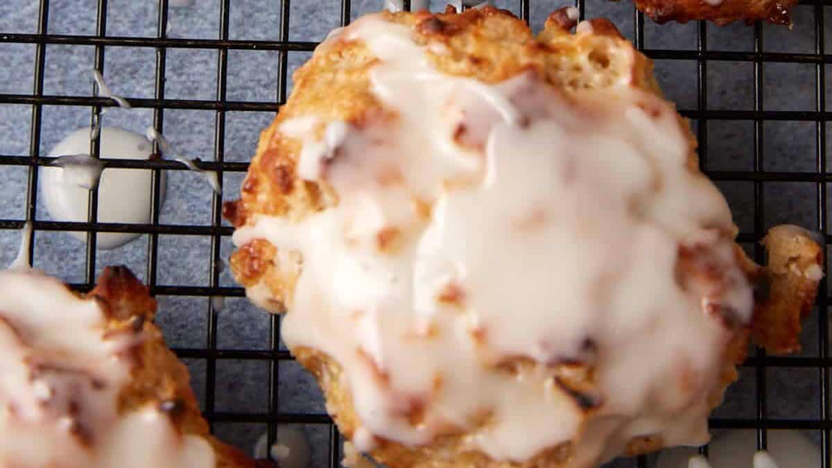 Four glazed scones on a black wire cooling rack. The scones are golden-brown with a thick white icing drizzled over the tops, partially covering the surface. The background shows parchment paper under the cooling rack.