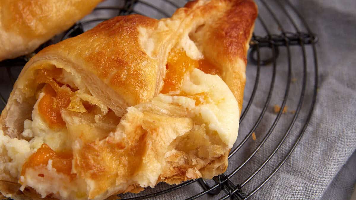 A flaky pastry filled with a creamy mixture and pieces of orange fruit, partially broken to reveal the filling, sits on a round cooling rack on a gray cloth. In the background, a bowl with more orange fruit pieces is slightly out of focus.