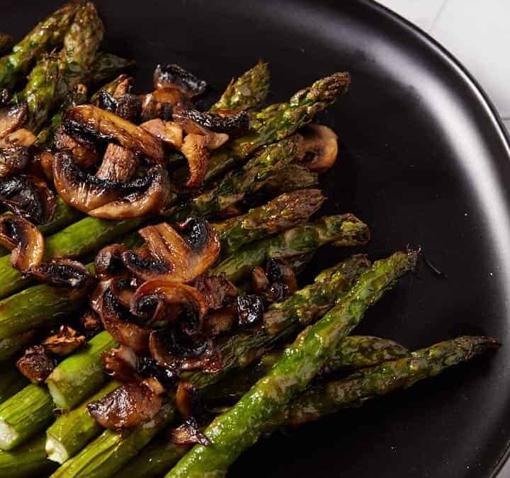 A platter of asparagus with mushrooms next to a container of balsamic vinegar