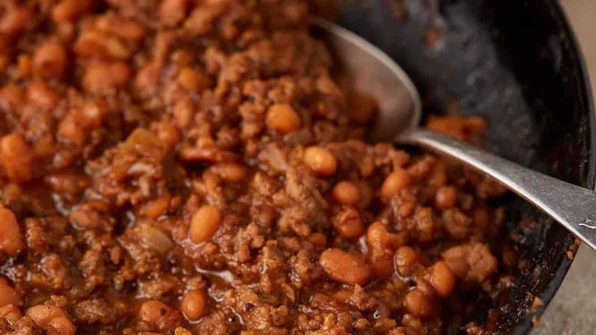 A skillet of baked beans with ground beef with a serving spoon.