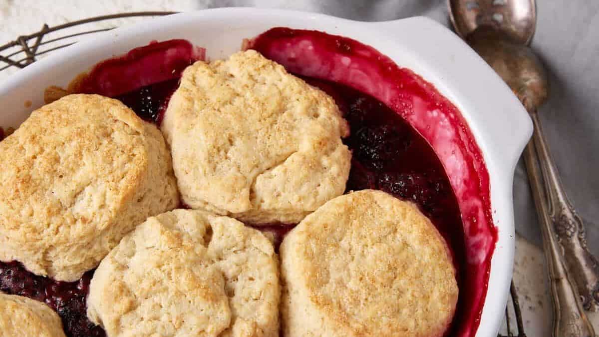 A white baking dish filled with freshly baked berry cobbler topped with six golden brown biscuits. The dish is placed on a cooling rack. Two spoons and stacked bowls, one pink and one green, sit in the background on a light-colored surface with a gray cloth—reminiscent of vintage recipes.