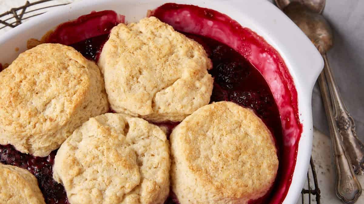 A white baking dish filled with freshly baked berry cobbler topped with six golden brown biscuits. The dish is placed on a cooling rack. Two spoons and stacked bowls, one pink and one green, sit in the background on a light-colored surface with a gray cloth—reminiscent of vintage recipes.