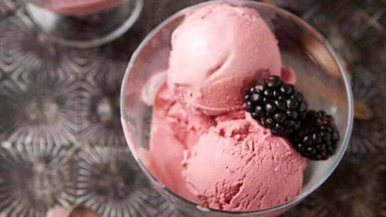Three glass bowls filled with scoops of blackberry ice cream topped with fresh blackberries are arranged on a textured metal surface. Two spoons, one with a small amount of ice cream, are placed beside the bowls.