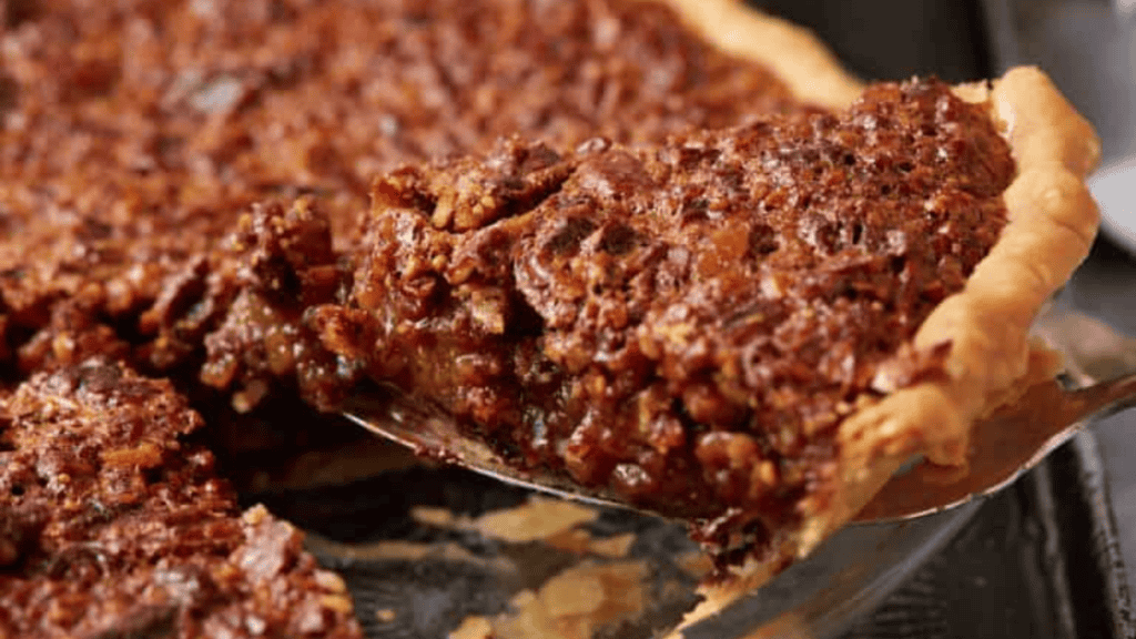 A close-up of a pecan pie with a golden crust, showing one slice being lifted out. The dense, caramelized filling packed with chopped pecans makes this classic an irresistible choice for easy desserts.
