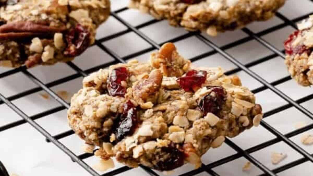 Close-up of several oatmeal cookies with visible nuts and dried cranberries cooling on a black wire rack. The cookies appear chunky and textured, with a mix of oats and dried fruit. The background is out of focus, highlighting the cookies.