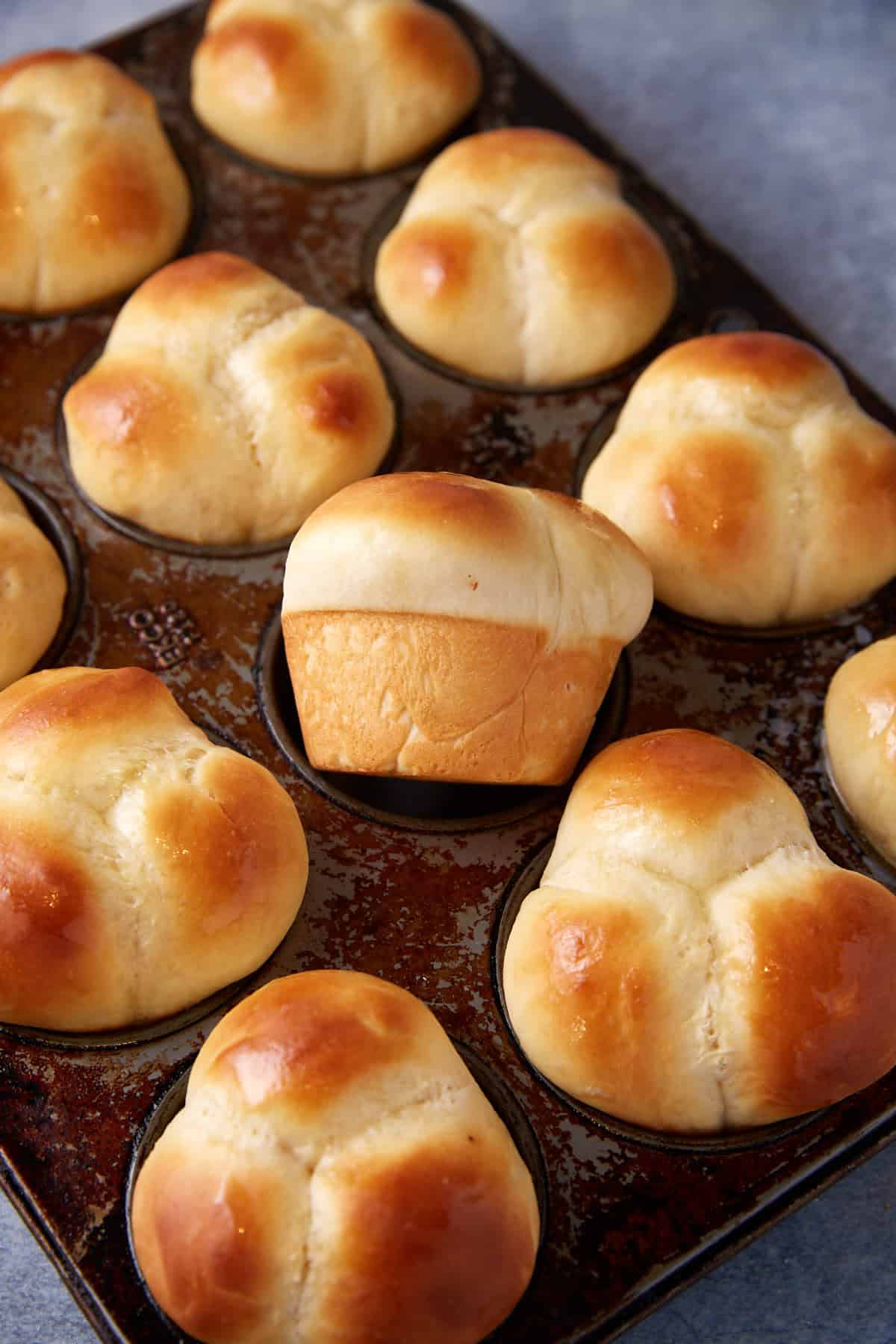 Freshly baked golden-brown cloverleaf dinner rolls are nestled in a muffin tin. One roll is slightly elevated, creating a whimsical, uneven appearance among the otherwise uniform set of rolls. The rolls are glossy and golden, indicating they have been brushed with butter.