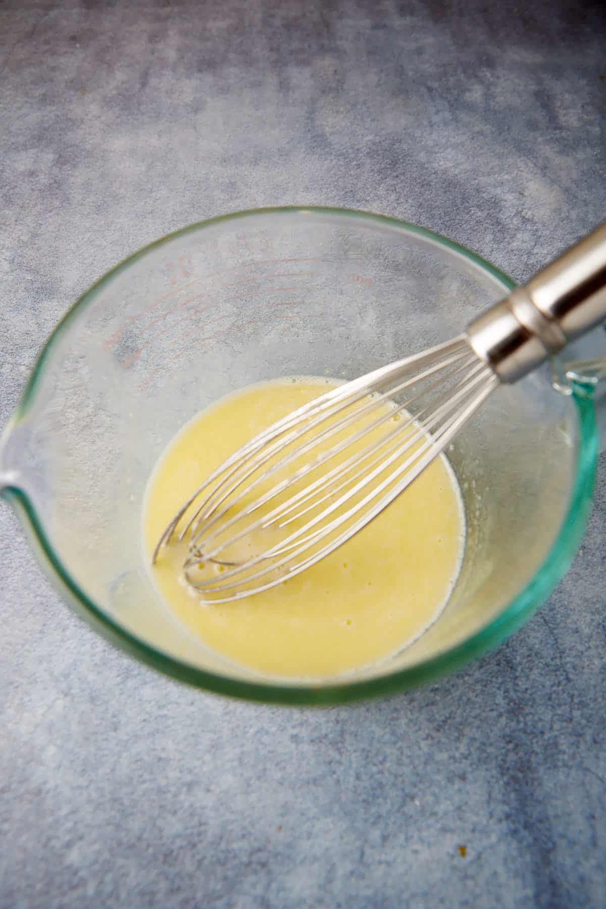 A glass mixing bowl containing a light yellow liquid is placed on a grey surface. A metal whisk is partially submerged in the liquid, indicating that mixing is in progress. The scene suggests preparation of a recipe, likely involving liquid ingredients.