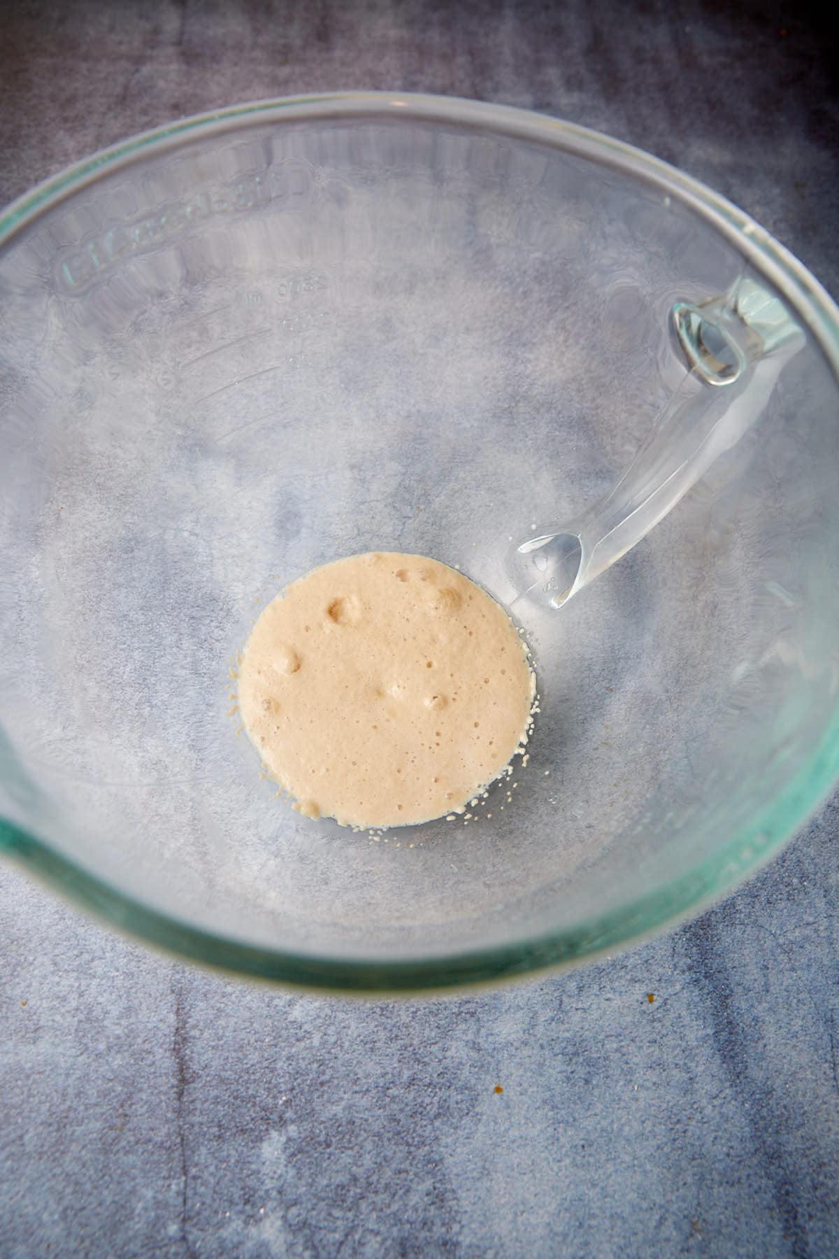 A clear glass mixing bowl on a gray countertop, containing a small amount of foamy, beige yeast mixture at the bottom.