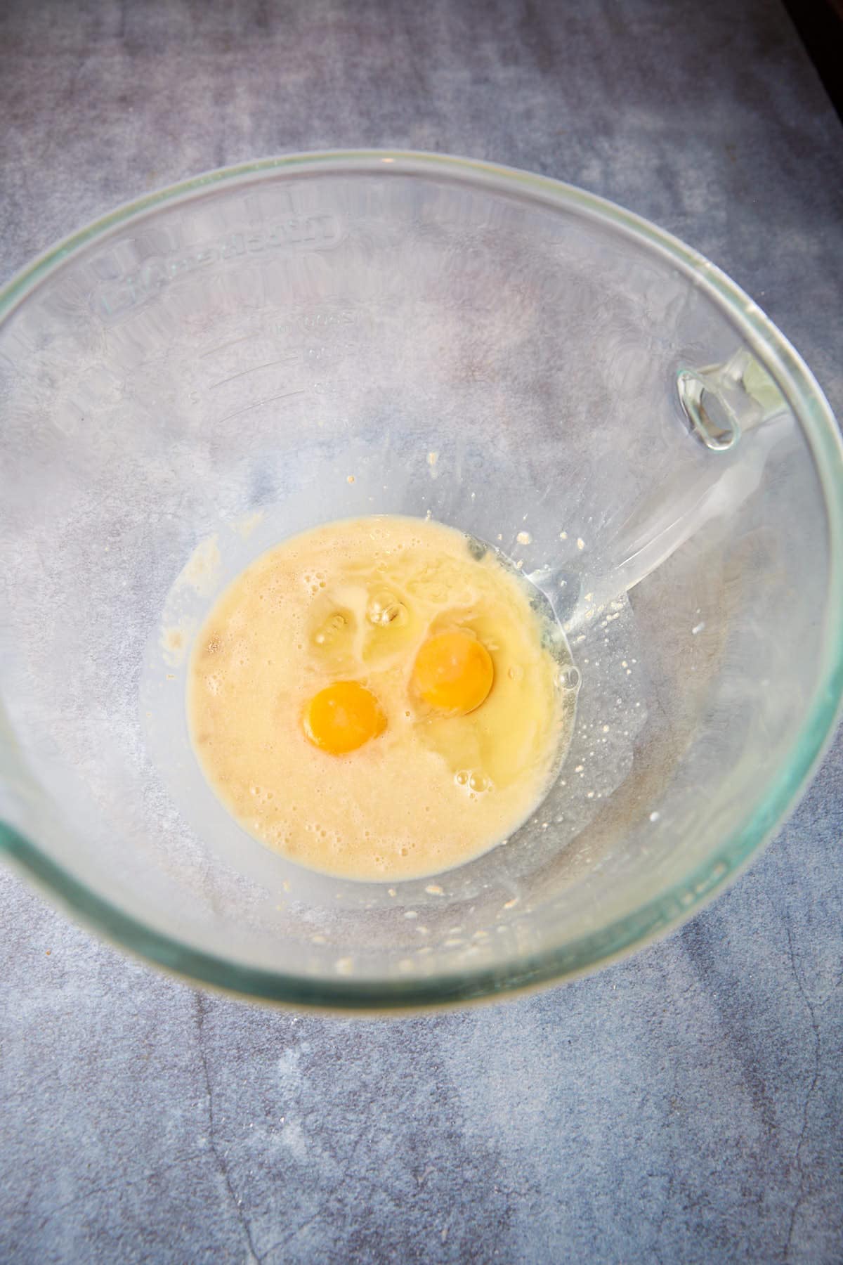 A glass mixing bowl sits on a gray countertop, containing a mixture of two cracked eggs with their yolks intact, partially blended into a light yellow liquid.