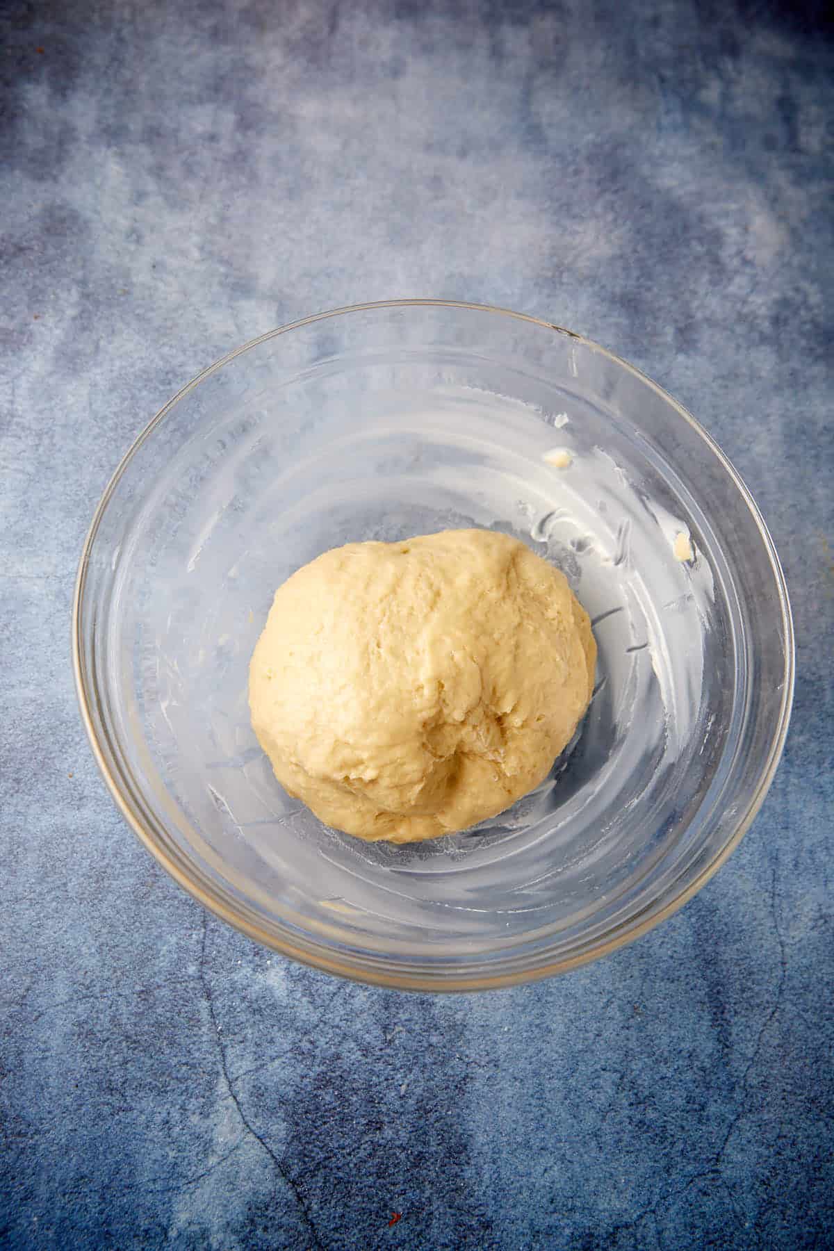 A clear glass bowl sits on a blue, textured surface, containing a ball of dough. The dough appears smooth and slightly moist, suggesting it has been kneaded and is ready for further preparation or rising. The background has a worn, rustic look.