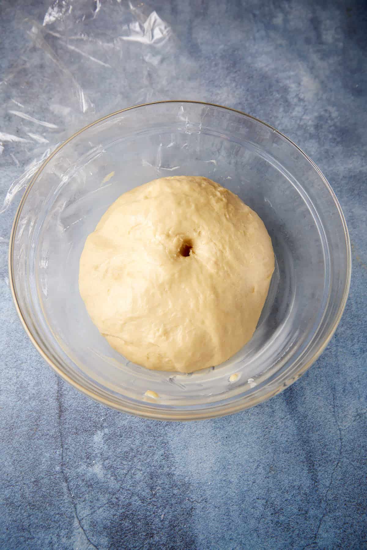 A ball of yeast dough rests in a clear glass bowl on a blue textured surface. The dough has a smooth surface with a small hole in the center, indicating it has been left to rise. Plastic wrap is partially visible at the top of the image.