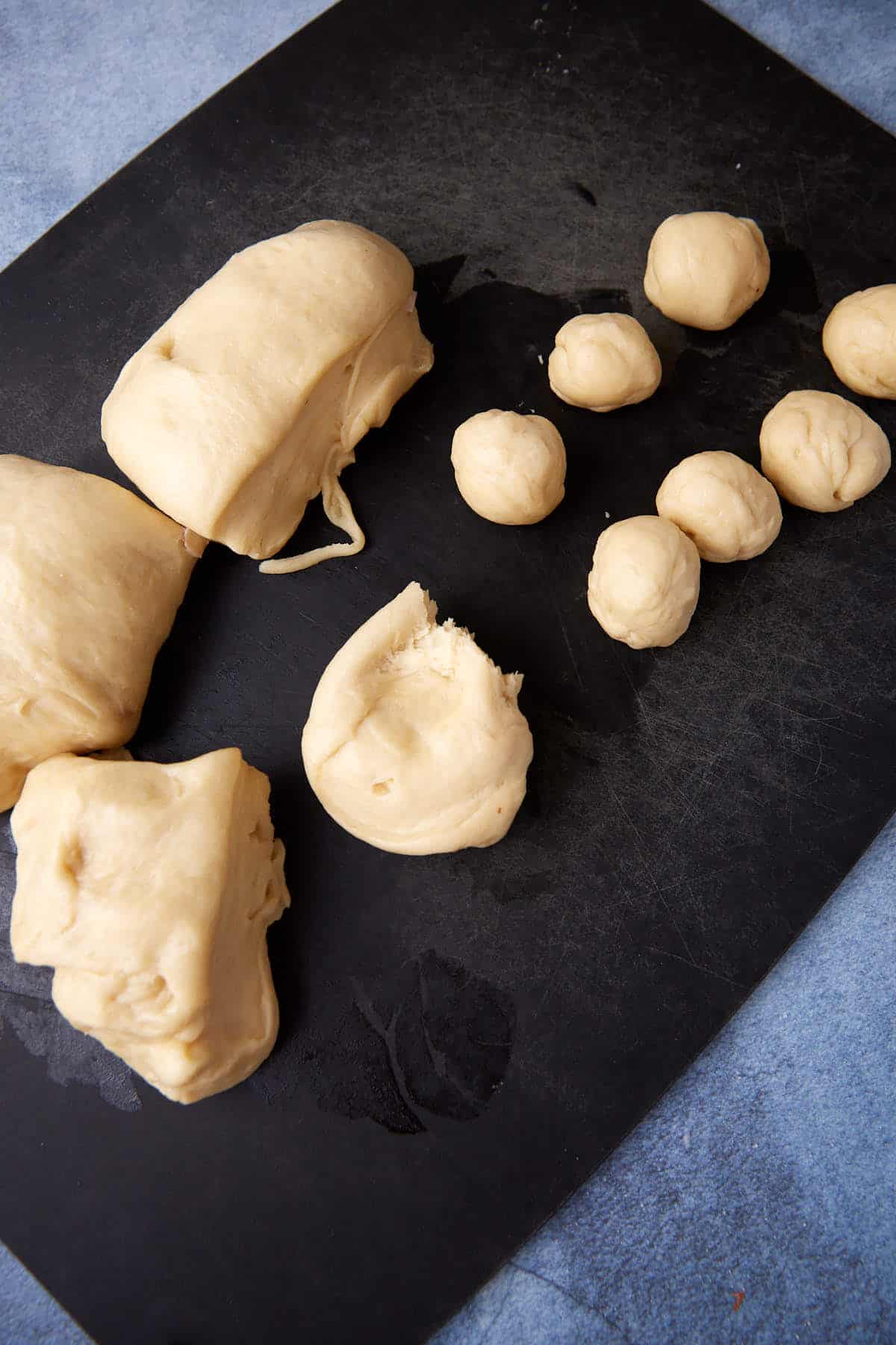 Several pieces of raw dough on a dark surface, with some formed into balls of varying sizes. The dough appears soft and slightly sticky, set against a blue background.