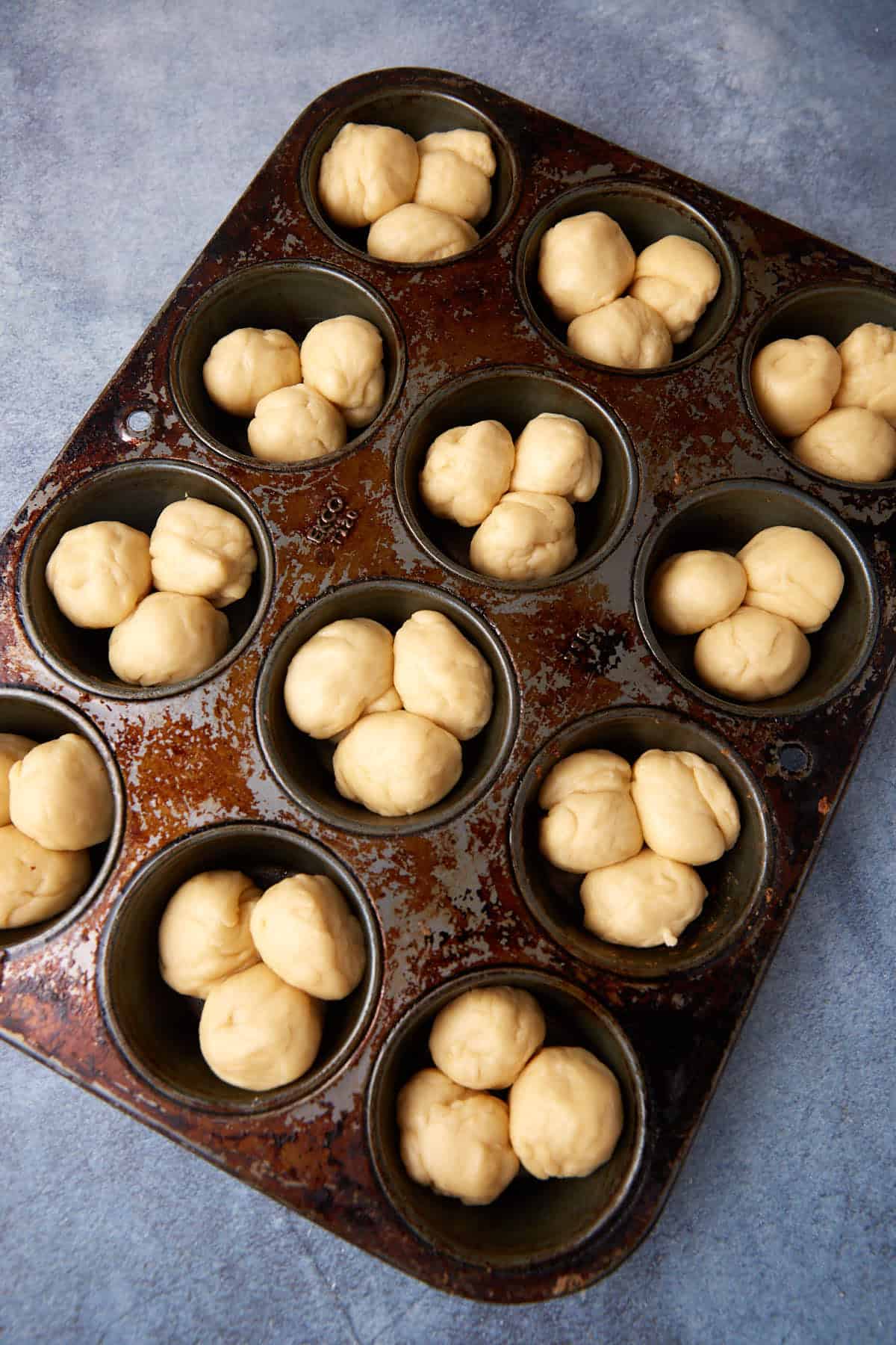 A muffin tin with twelve slots is filled with clusters of small dough balls, ready for baking. The tin shows signs of wear, and the background is a blue-gray surface.