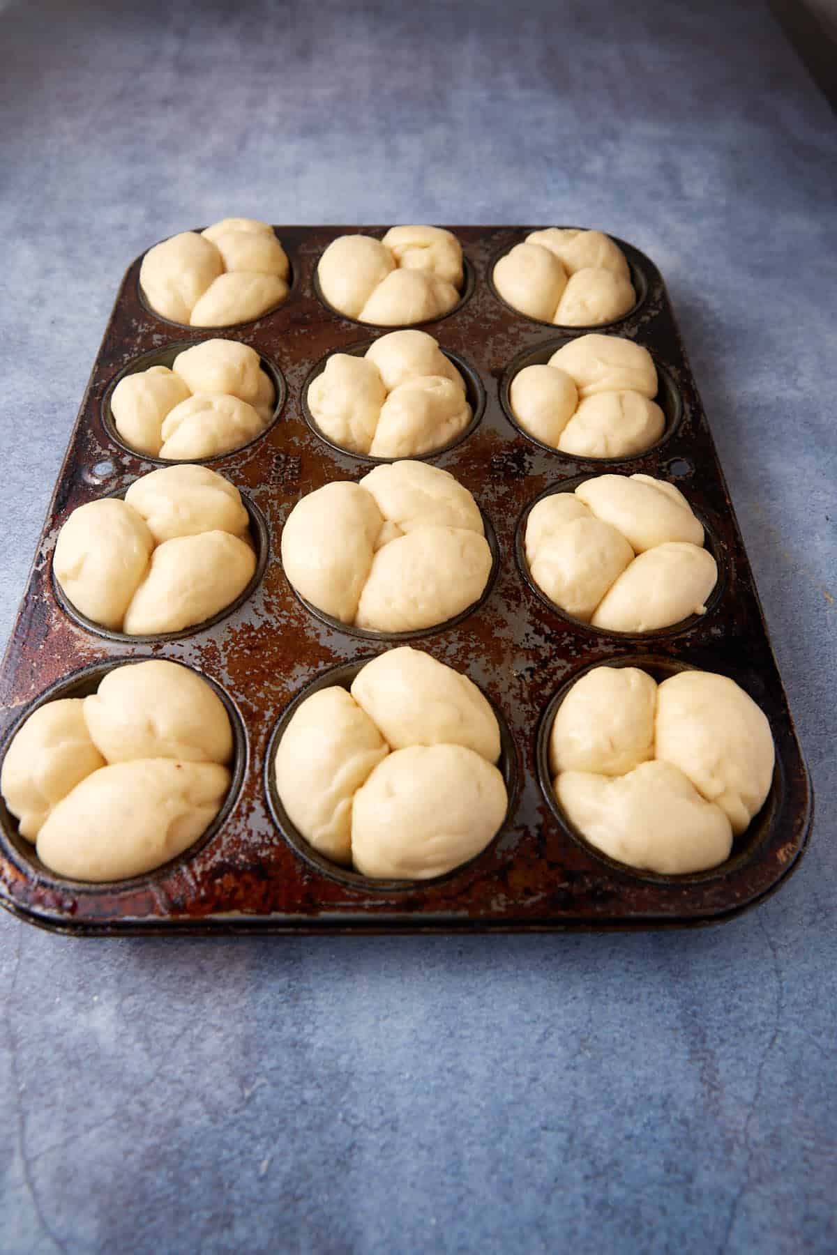A dark metal muffin tray with twelve compartments is filled with pre-baked cloverleaf dinner roll dough divided into three segments per compartment. The tray rests on a light blue textured surface, ready to be baked.