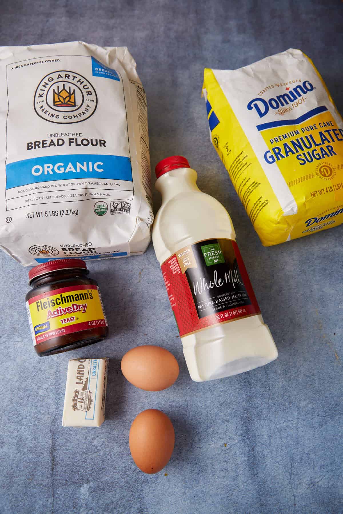 A selection of baking ingredients is arranged on a blue surface, perfect for making brioche rolls: a bag of organic bread flour, a bag of granulated sugar, a bottle of whole milk, a jar of active dry yeast, two eggs, and a stick of unsalted butter.