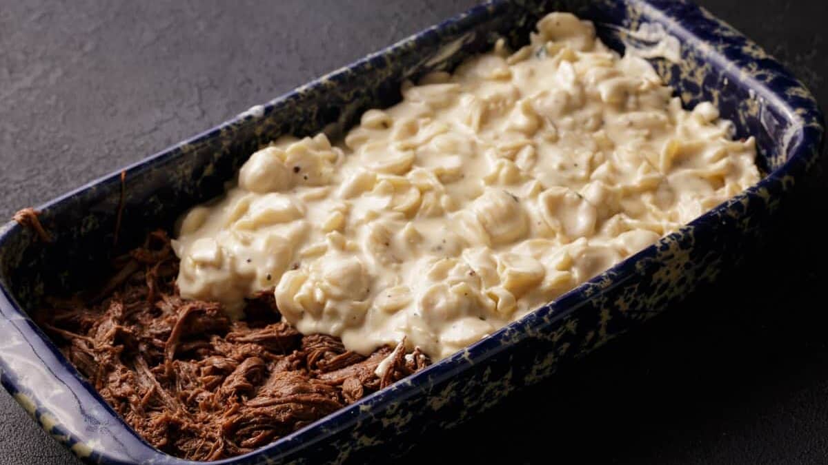 A casserole dish with brisket on the bottom and half covered with pasta.