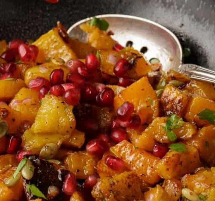 A bowl of roasted butternut squash salad with pomegranates being sprinkled on top.