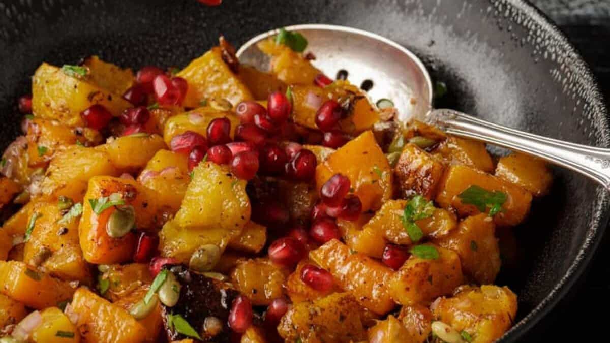 A bowl of roasted butternut squash salad with pomegranates being sprinkled on top.