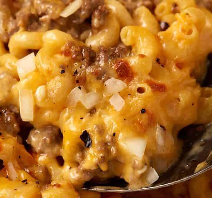 A close-up image of a skillet filled with creamy cheeseburger macaroni mixed with ground beef and topped with chopped onions. A serving spoon is lifting a portion of the cheesy dish, showcasing the melted cheese and rich texture.